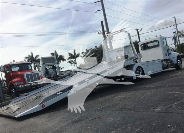 Peterbilt 337 tow truck with a heavy-duty flatbed in a dealership lot; white cab, long wheelbase, other trucks visible in background.