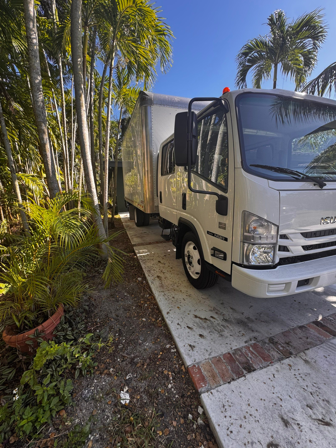 Isuzu NQR box truck exterior with cargo box, front grille showing Isuzu emblem, side mirror, and step; tropical plants and driveway around.