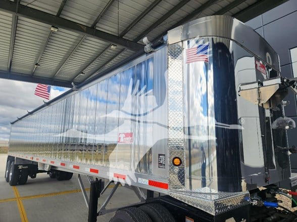 A shiny chrome-trimmed dump trailer with side panels reflecting the sky; features include American flags, reflective safety chevron strips, and a front-loading mechanism. Visible logo on side reads 'Travis' trailer and there is a small red