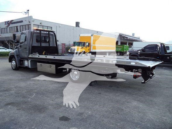 Tow truck with a flatbed in a lot; no legible labels or fluid specs visible on the vehicle in the foreground.