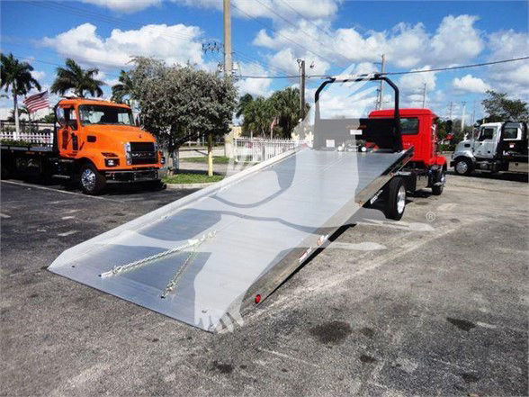 Flatbed tow truck bed with bright aluminum surface and ramp, red truck cab in background, blue sky with some clouds.