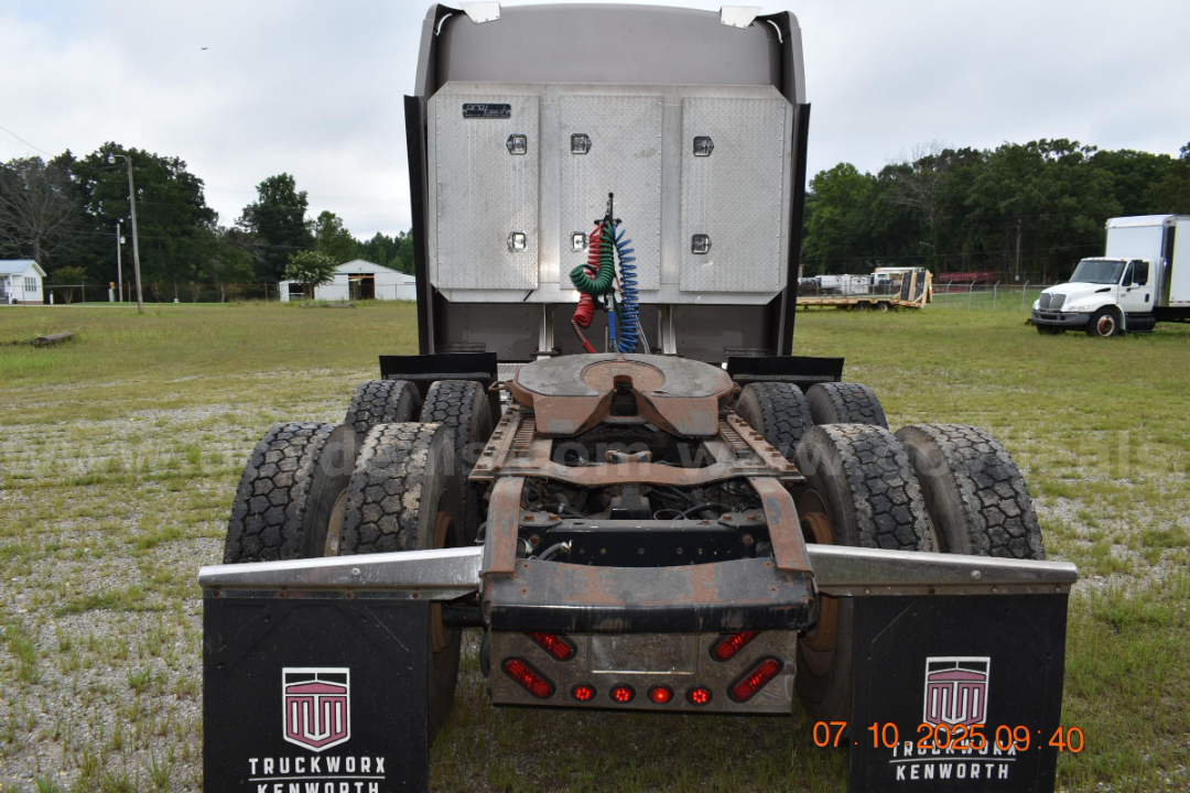 Kenworth W900 cab and chassis rear view showing fifth wheel, dual rear axles, and air lines; metal diamond-plate toolbox visible behind cab; dirt and rust on chassis.