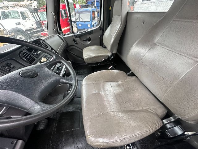 Truck interior cab with steering wheel, dashboard, and two bucket seats; worn fabric on seats; view through windshield shows other trucks outside.