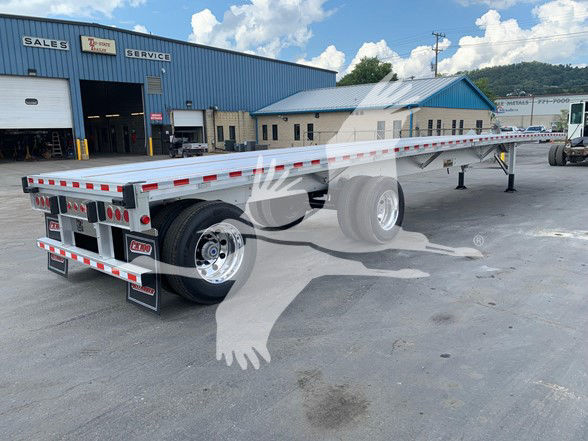 Flatbed trailer on a dealership lot with dual rear axles and chrome wheels, long platform with reflective tape along sides.
