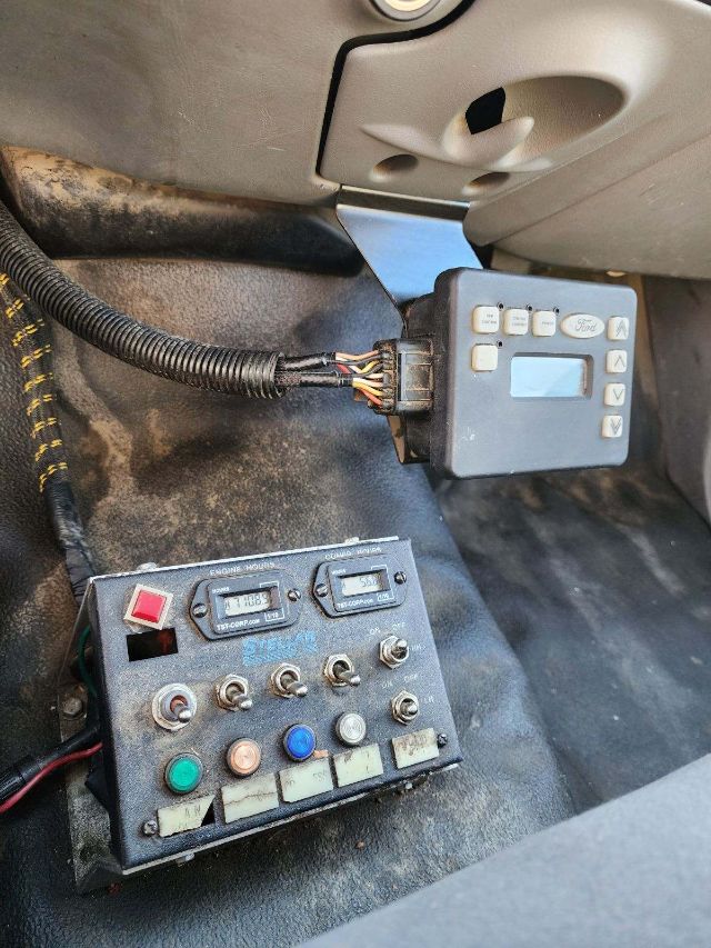 Truck dashboard area showing a blue control panel with multiple buttons, a small display, and a wired connector; another metal control module with colored push buttons and switches on a dirty floor surface.
