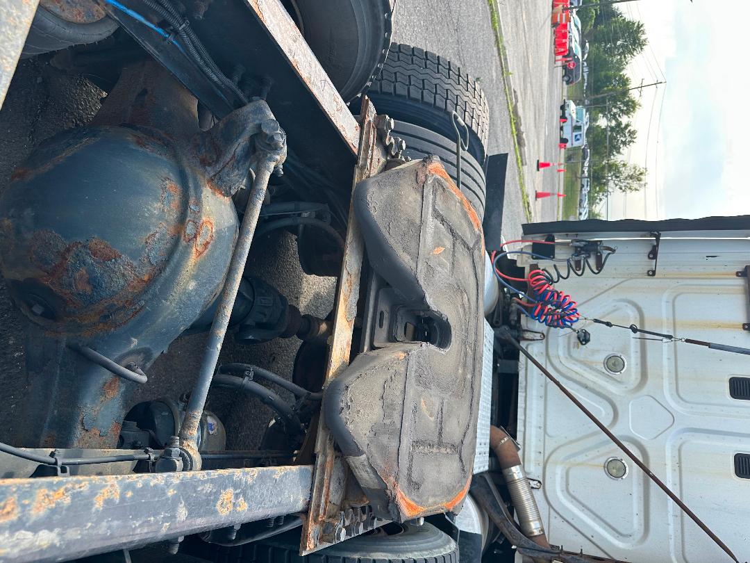 Freightliner Cascadia underside showing drive axle housing, suspension components, and spare wheel on trailer; rusted metal and worn bumper area visible.