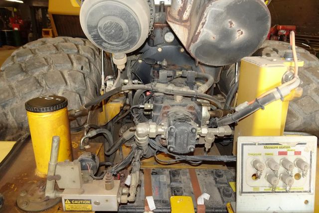 VM106D Vibromax truck engine bay showing hydraulic pump, hoses, reservoirs, and control panel with labels and gauges.