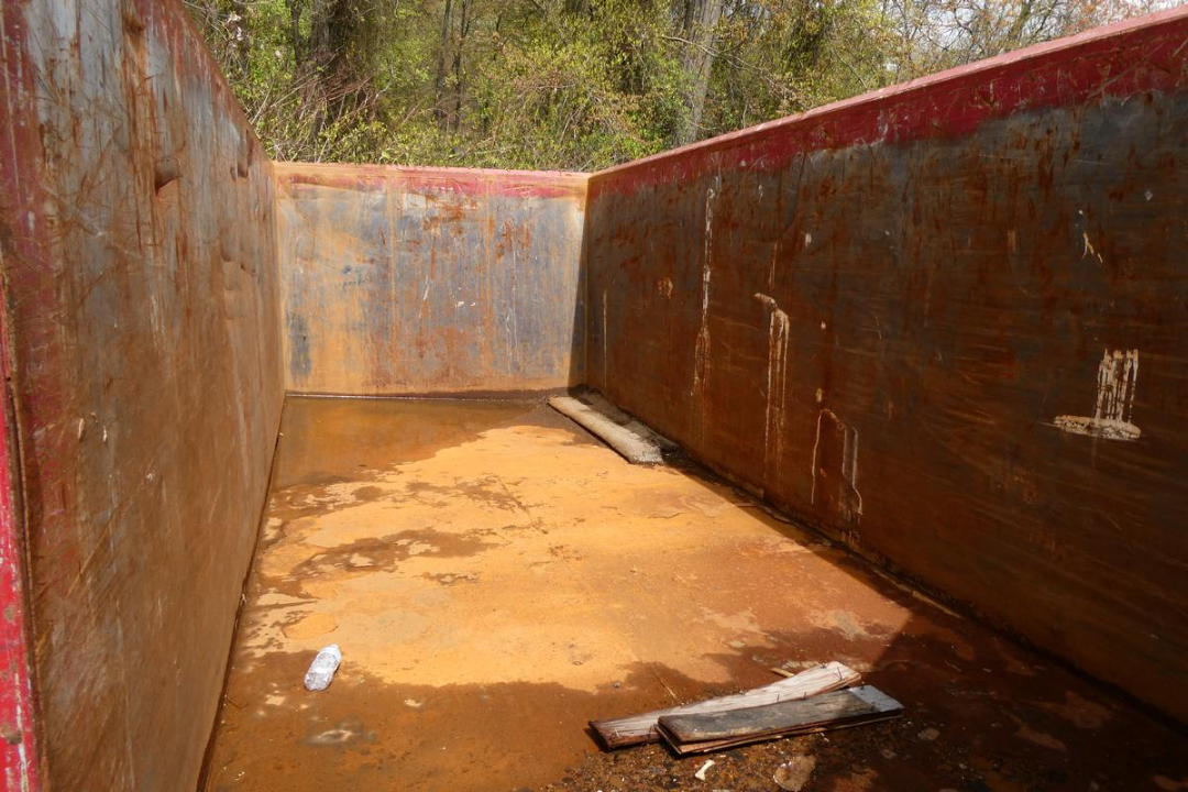 Inside a rust-stained empty dump truck bed with wooden sidewalls and scattered debris including a water bottle.