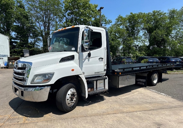 20120 Hino 258 tow truck flatbed in a parking lot with green trees in background; white cab and black flatbed; VIN visible on listing.