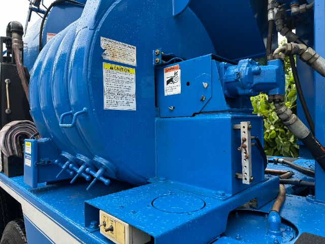 Blue Mack GU713 vacuum truck rear/side with warning labels, hoses, and hydraulic components. Visible text includes caution labels on the tank and control housing.
