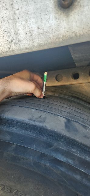 Wheel/tire mounted inside trailer frame with a hand using a green-titted tool near a metal beam; visible tire tread and inner wheel area.