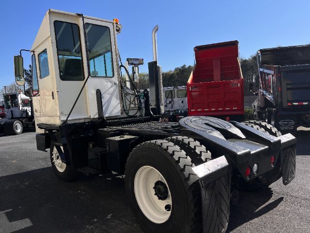 Vehicle chassis with white cab and black wheel arches, Kalmar Ottawa heavy truck tractor in yard, red dump body in background.