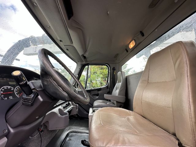 Truck interior driver area of a 2011 Freightliner M2 with tan seats, steering wheel, dashboard, and manual gear shifter.