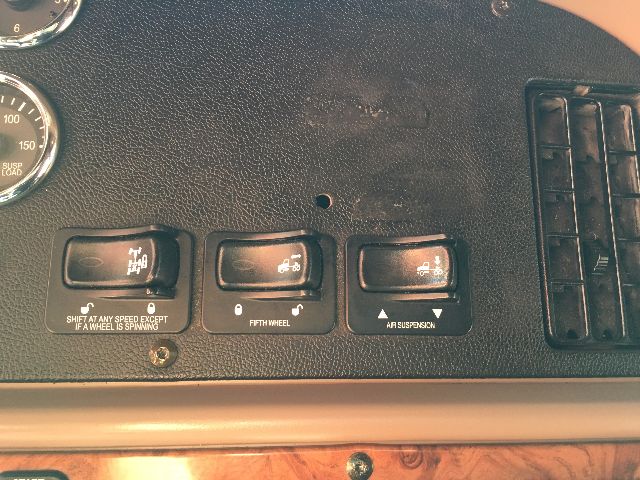 Dashboard switches and gauges in a Peterbilt truck console showing three rocker switches with white icons; text under switches reads 'SHIFT AROUND SPEED EXCEPT IF A WHEEL IS SPINNING', 'FIFTH WHEEL', and 'AIR SUSPENSION'. A gauge/needle is