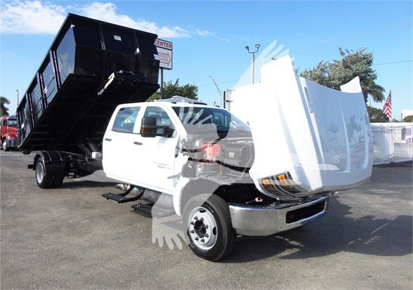 2023 Chevrolet Silverado 5500HD dump truck in a dealership lot, white cab, large black dump bed raised behind cab, front hood open.