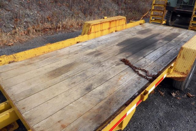 Trailer platform showing weathered wooden deck with a chain, yellow steel frame, and wheel fender.