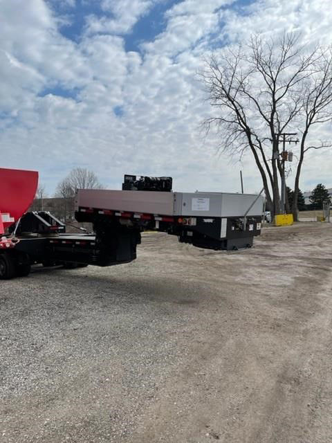 Semi-trailer trailer with flatbed deck and black hydraulic lift mechanism, side rails, and a gray upper deck. Clear sky with trees in background.
