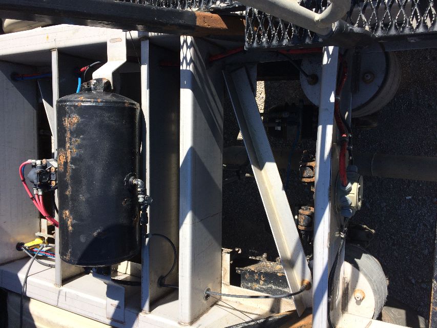 Pneumatic dry bulk trailer area showing a black cylindrical air tank mounted on a white frame with hoses and wiring around; visible wheel and axle components.