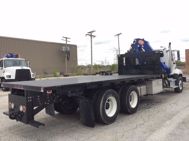 Flatbed truck with knuckle boom crane mounted near rear, white cab, blue crane, black metal flatbed, triple rear axles. Visible on truck: blue knuckle boom crane near front of flatbed.