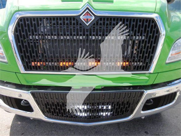 Front view of a green 2024 International MV truck showing the grille and bumper, with a translucent overlay of dove-like shapes.