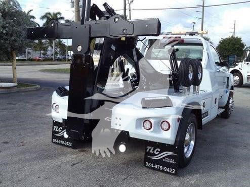 Tow truck with a large black tow crane and platform on white Chevy Silverado 6500HD, rear view showing stabilizers and equipment, TLC contact numbers visible on mudflaps.