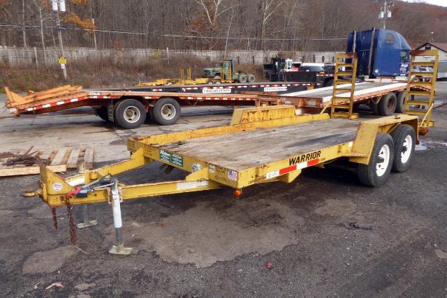 Yellow Warrior brand tilt trailer with wood deck and dual axles, showing 'WARRIOR' on the side and a hitch at the front; there are other trailers and trucks in the background.