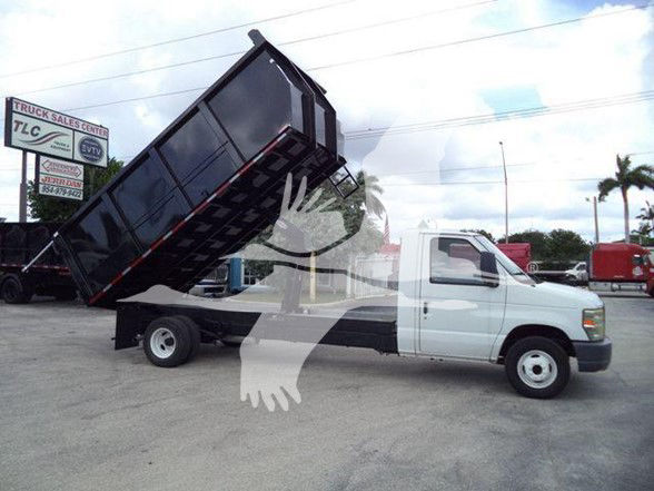 2011 Ford E450 dump truck with black dump bed raised at an angle, white cab, stock yard in background. Visible text on a sign in the lot: TLC Truck Sales Center (partial).