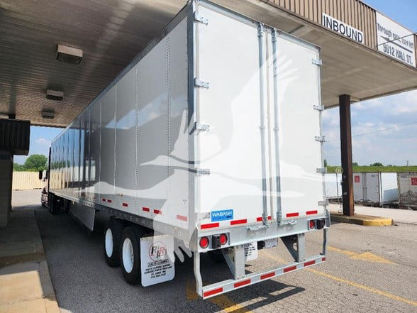 Large white Wabash trailer in a loading dock area, rear view showing double doors, taillights, and red/white reflective stripes. Wabash logo near bottom rear.