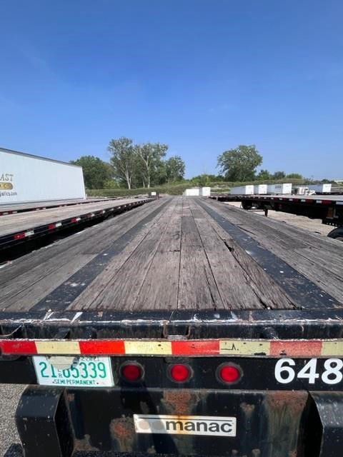 Flatbed trailer bed with wooden planks and Manac branding, rear view showing tail lights and Minnesota license plate.