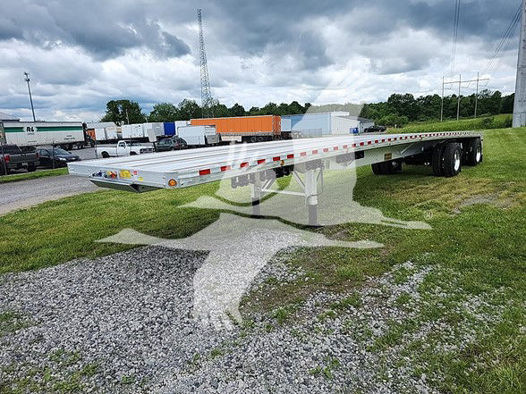 Flatbed trailer shown from side/front angle with tandem axles in background and reflective tape along the edge.