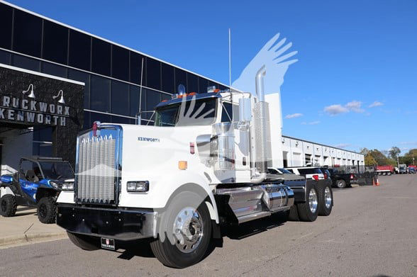 Kenworth W900L conventional daycab truck in front of dealership with chrome exhaust stacks and large grille.