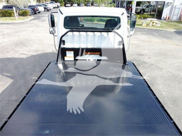 Flatbed truck bed with a Freightliner Business Class M2 106 cab in the background. The metal diamond-plate flatbed shows a shadowy reflection.
