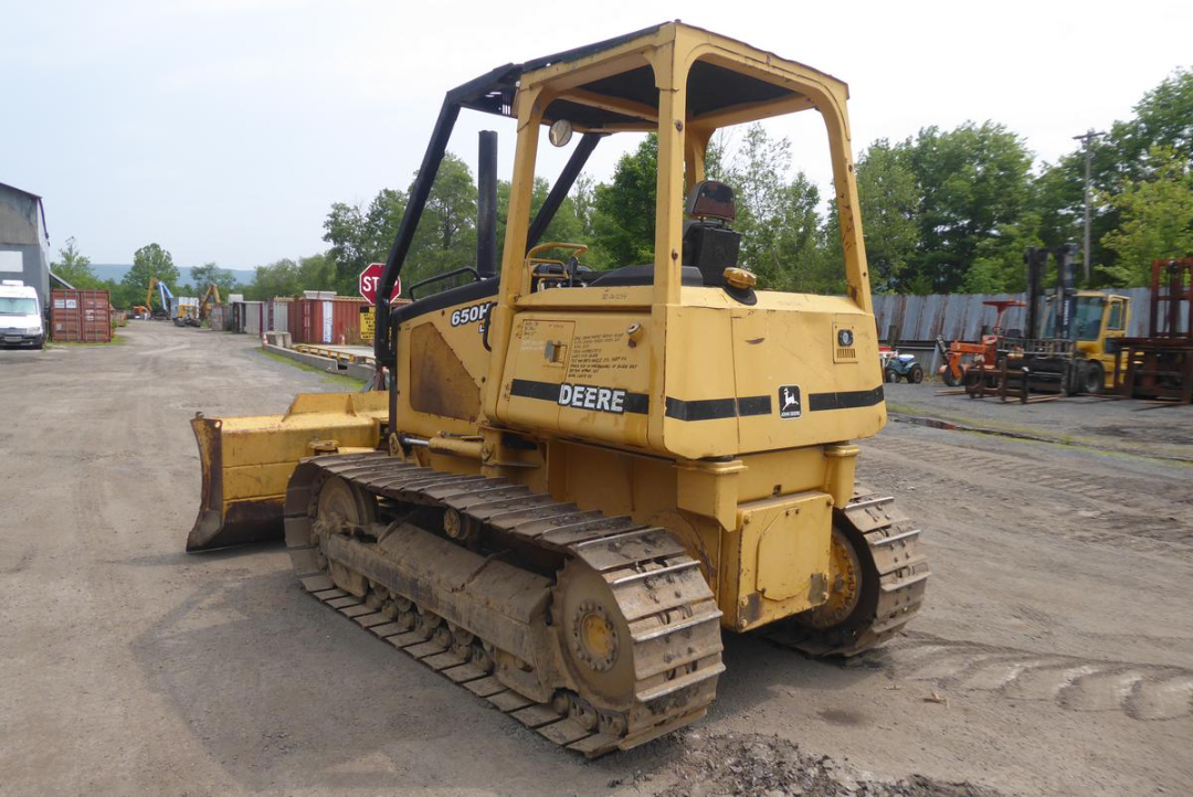 John Deere 650HLT tracked bulldozer with front blade, yellow paint, operating in a lot with other equipment in background.