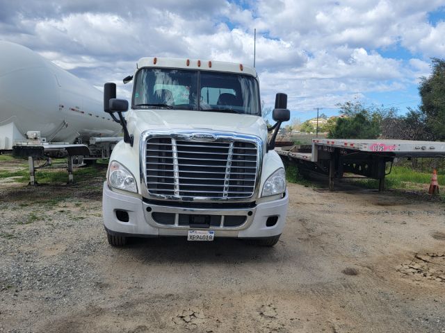Freightliner Cascadia front view with chrome grille, large headlights, white cab. License plate visible on bumper, antennae on roof, flatbed trailer to right and tanker trailer to left.