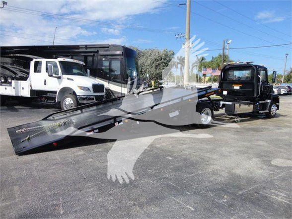 Tow truck with a long flatbed ramp in a dealership lot. A white truck is visible on the left, a black tow truck on the right, and a large gray ramp extending toward the foreground.