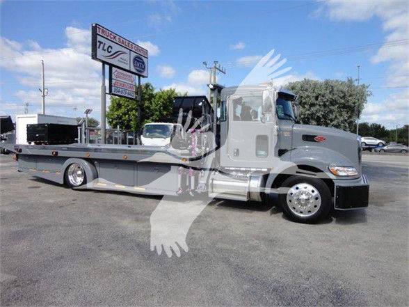Truck with a long flatbed, Peterbilt badge on the cab, side mirrors, and a watermark overlay. Visible text includes TLC Truck Sales Center on a sign in the background. The cab shows Peterbilt styling with a prominent grille.