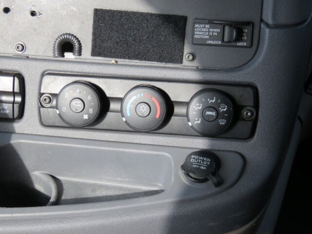 Dashboard of a Freightliner Cascadia Evolution showing climate control knobs (left: fan and temp, center: temp dial with blue/red, right: defrost/airflow), a 12V power outlet cap labeled POWER OUTLET, and a small panel with a note 'MUST BE 