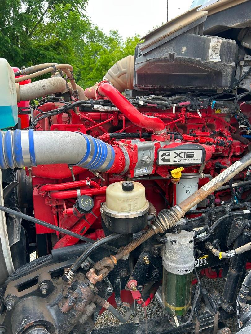 Kenworth X15 engine bay showing red engine block, blue intake hose, coolant reservoir, and various hoses and cables visible.