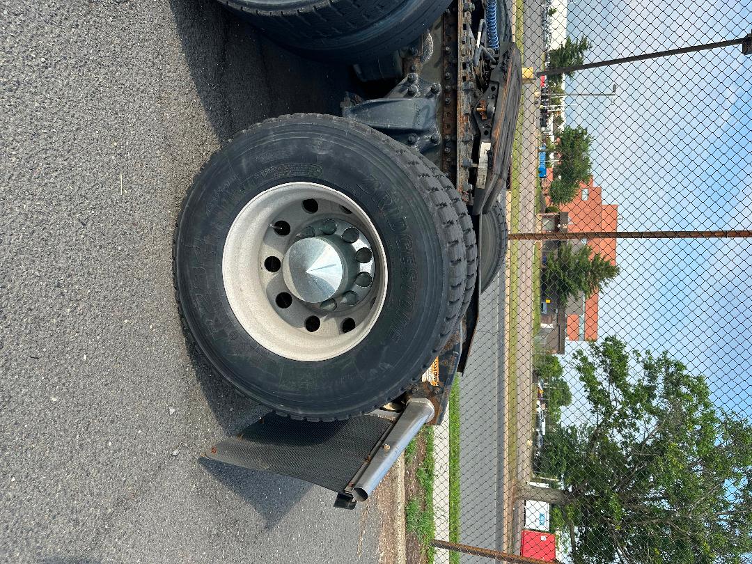 Truck wheel/tire assembly with dual rear wheels and metal hubcap; visible chains and chassis components under Freightliner Cascadia trailer, outdoor lot.