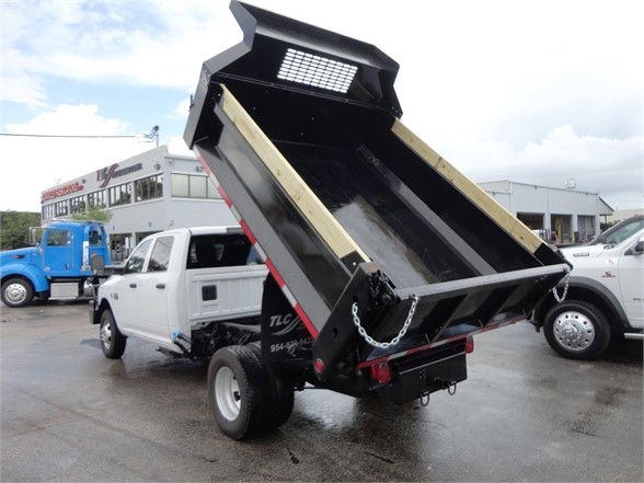 Dump truck tailgate raised showing empty bed and chain, Ram 3500 in background; several trucks in a dealership lot.
