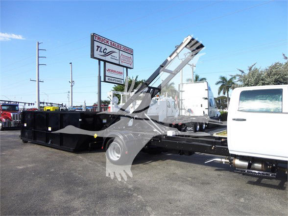 Truck with a large flatbed and a hydraulic crane/crane arm in a parking lot. Visible signs in background read 'TRUCK SALES CENTER' and other indistinct text on a billboard.