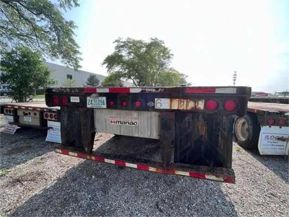 Manac flatbed trailer rear view with rusted frame, round taillights, and rear bumper; multiple license plates and a Manac logo plate visible.