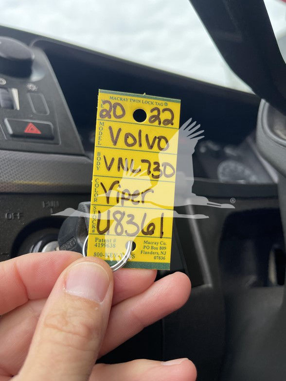 Tag with handwritten text: 'VOLVO VNL 73 0' and numbers 20 22, U8361, with other small text on tag. Background shows dashboard area.