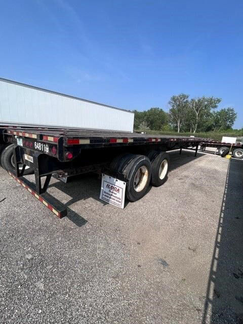 Flatbed trailer with dual rear axles and Manac branding. Visible are rear lights, license plate area, and a small sign on the ground. Text visible includes numbers on the trailer and a sign reading something like ‘KQCA’ or dealer plate.