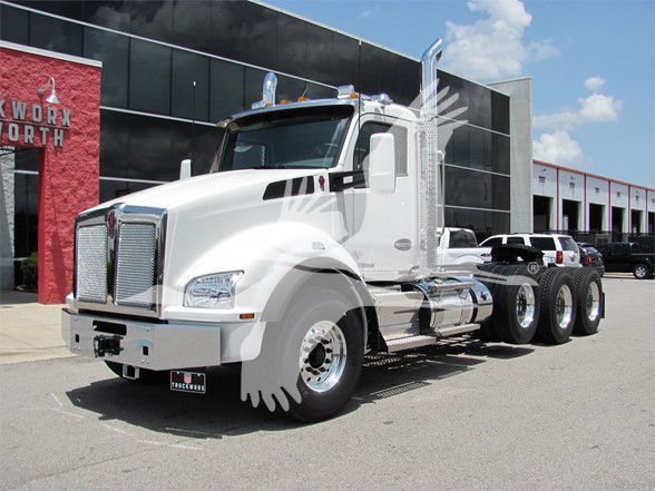 Kenworth T880 conventional daycab truck in white with chrome accents, large grille, dual stacked exhaust stacks, three rear axles, dealership in background.