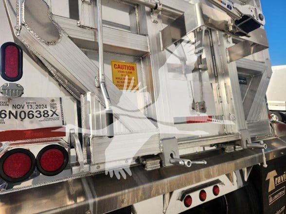 Back of a shiny metal dump trailer showing CAUTION label, taillights, and serial/registration plate. Visible text includes CAUTION, NOV 13, 2024 (on plate), and stock/lot details blurred.