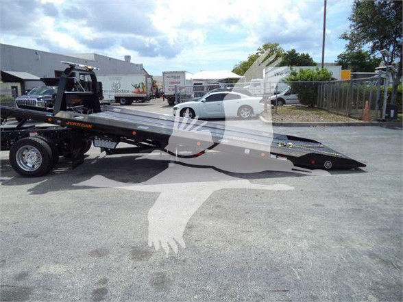 Ford F550 tow truck with a car hauler bed, trailer visible in background, dealership lot with other vehicles.
