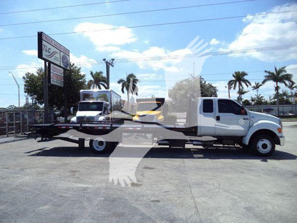 2015 Ford F650 XLT tow truck flatbed being towed by another truck; side view showing cab and long flatbed.