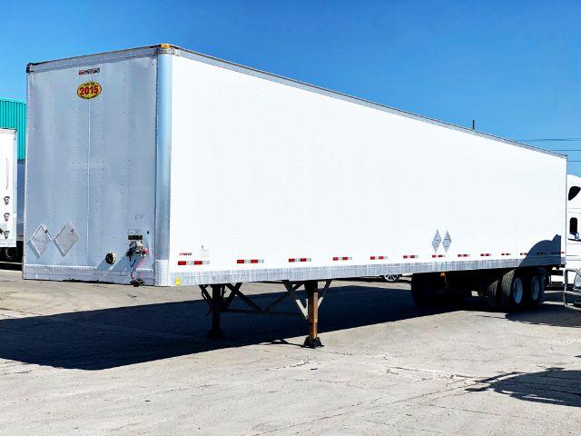 2015 Manac dry van trailer with white sides, front left corner shows a small oval '2015' sticker. Trailer parked on lot, support legs down, double rear wheels.