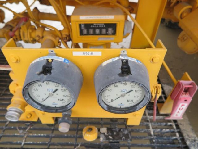 Two large round gauges labeled XSEL on yellow industrial control panel with a small digital display above showing gallons; label B/2018 attached; various pipes and valves around; pink pull handle on right.